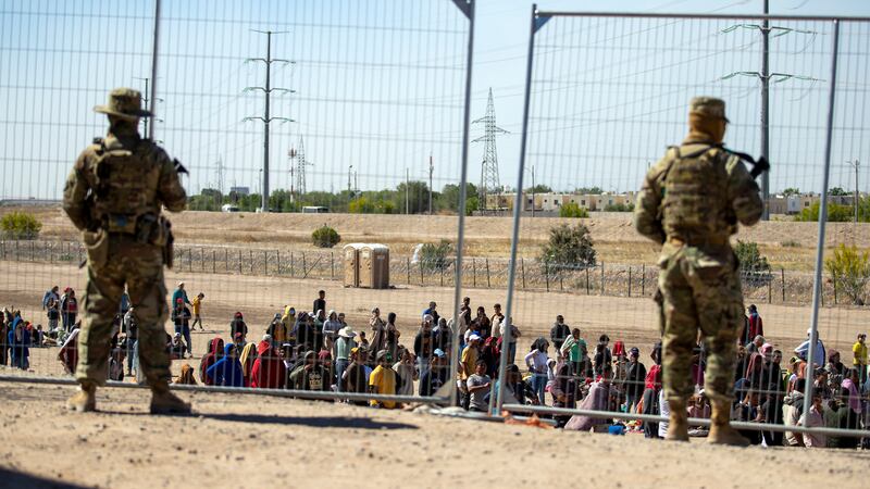 ARCHIVO - Varios migrantes esperan junto a la cerca fronteriza ante la mirada de guardias...